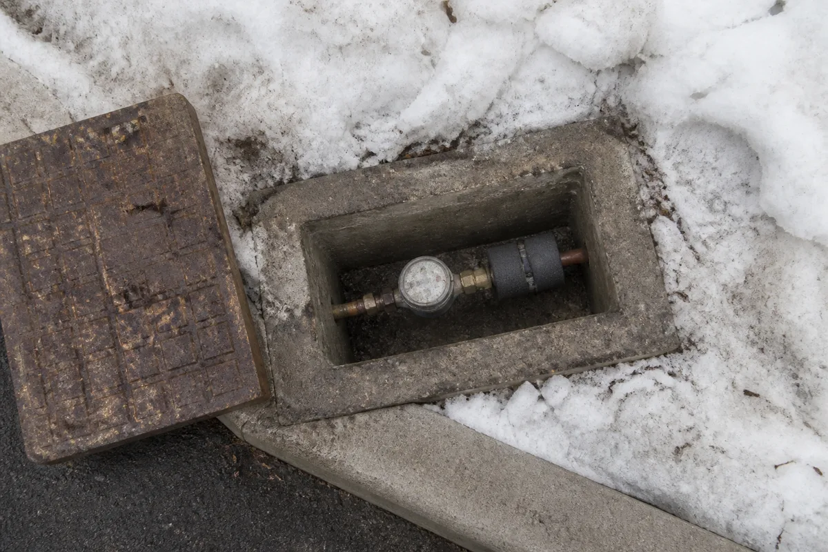 Regard de compteur d&rsquo;eau ouvert dans la neige au bord d&rsquo;une allée, isolant en mousse visible autour du tube