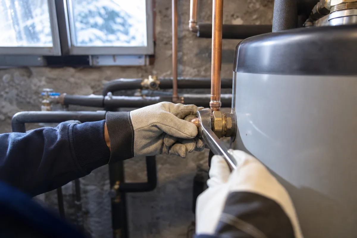 Mains d’un plombier resserrant un raccord de cuivre sur un chauffe-eau, lumière froide d’un vasistas de sous-sol