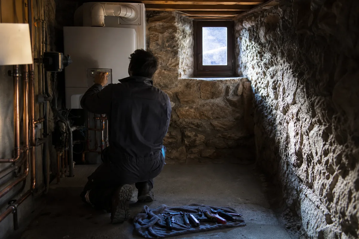 Technicien de dos réglant une chaudière murale dans une chaufferie de chalet savoyard, lumière froide de fin de journée